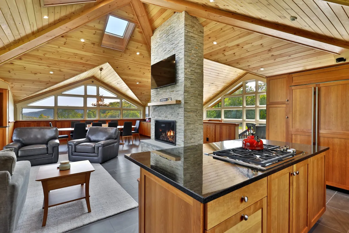 A central stone fireplace anchors this inviting open-plan space, surrounded by a comfortable lounge area and a kitchen with a prominent island. Large windows provide sweeping valley and ski trail views, while rich wood accents infuse the room with warmth.