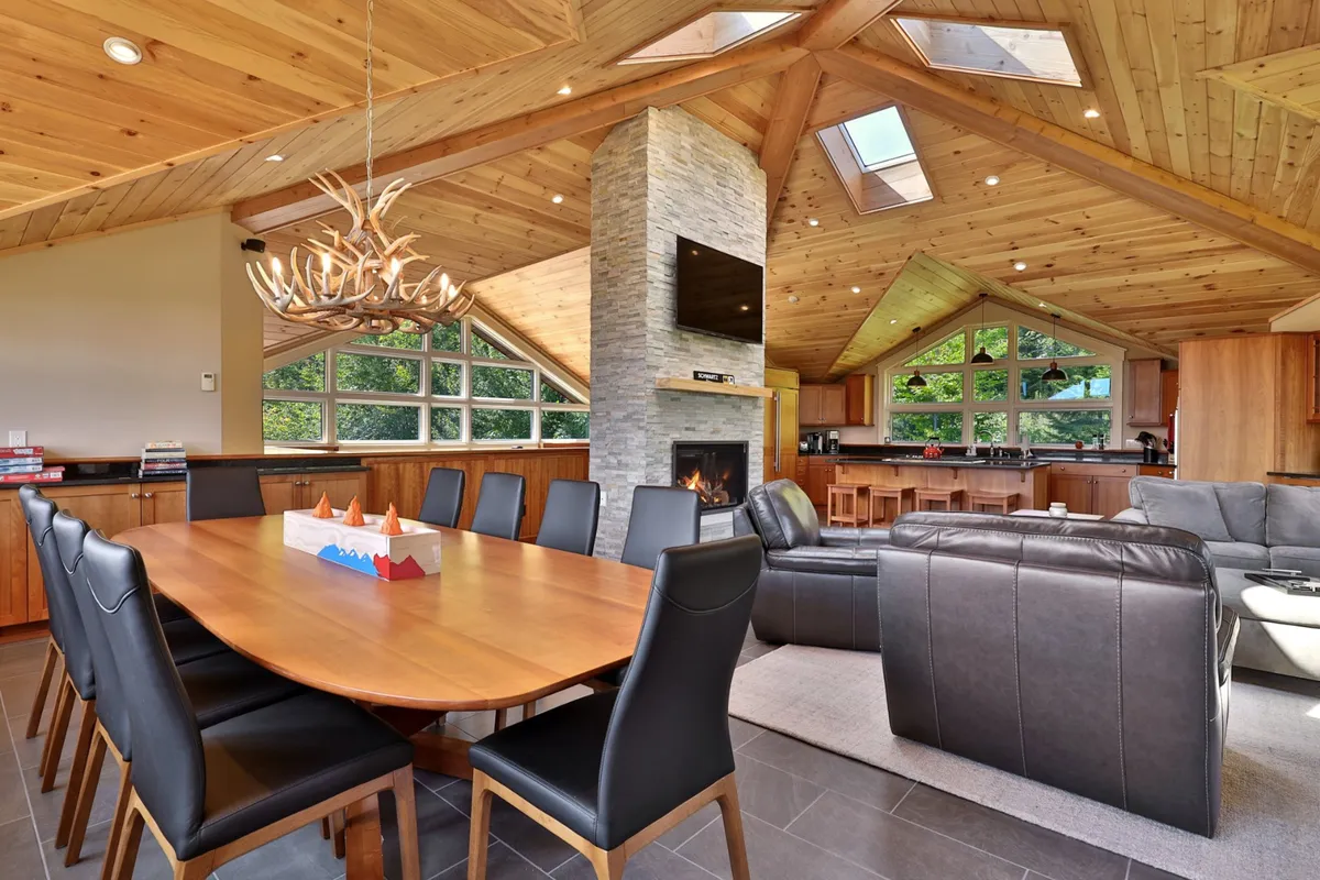 The image shows a stunning open-concept dining and living area framed by expansive windows that reveal breathtaking mountain views. A crackling fireplace sits at the heart of the space, casting a warm glow over a stylish dining table set for gatherings, while a grand chandelier adds a touch of mountain elegance. The scene exudes comfort and sophistication, perfect for memorable group moments after a day on the slopes.