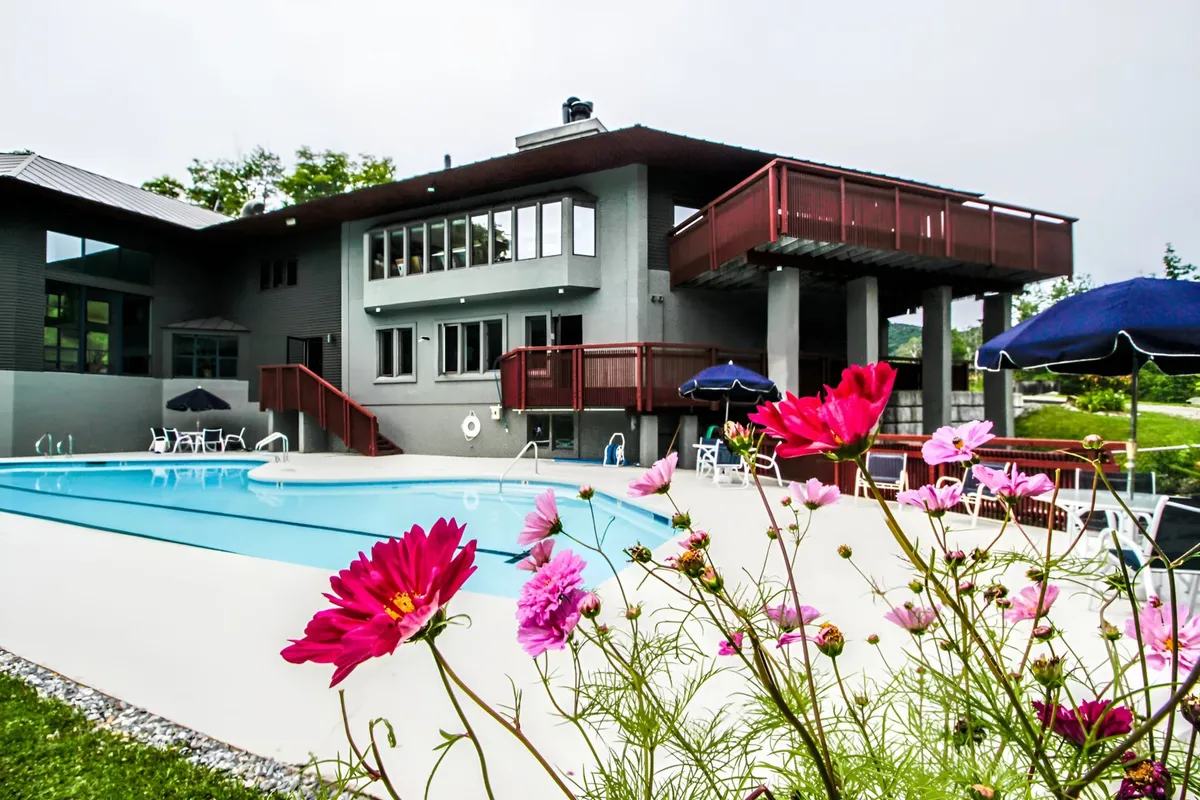 This exterior image features a sparkling swimming pool surrounded by flower beds, a deck area, and the house's windows in the background. The setting appears vibrant, inviting, and perfectly suited for family gatherings or relaxing after a day of adventure.