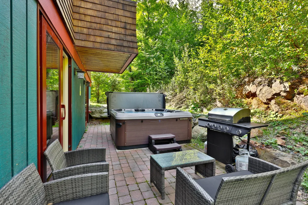 A welcoming outdoor area featuring a private hot tub, grill, and comfortable seating set against a tranquil stone wall. The space is ideal for après-ski relaxation and socializing, with the setting sun hinting at cozy evenings after a day on the slopes.