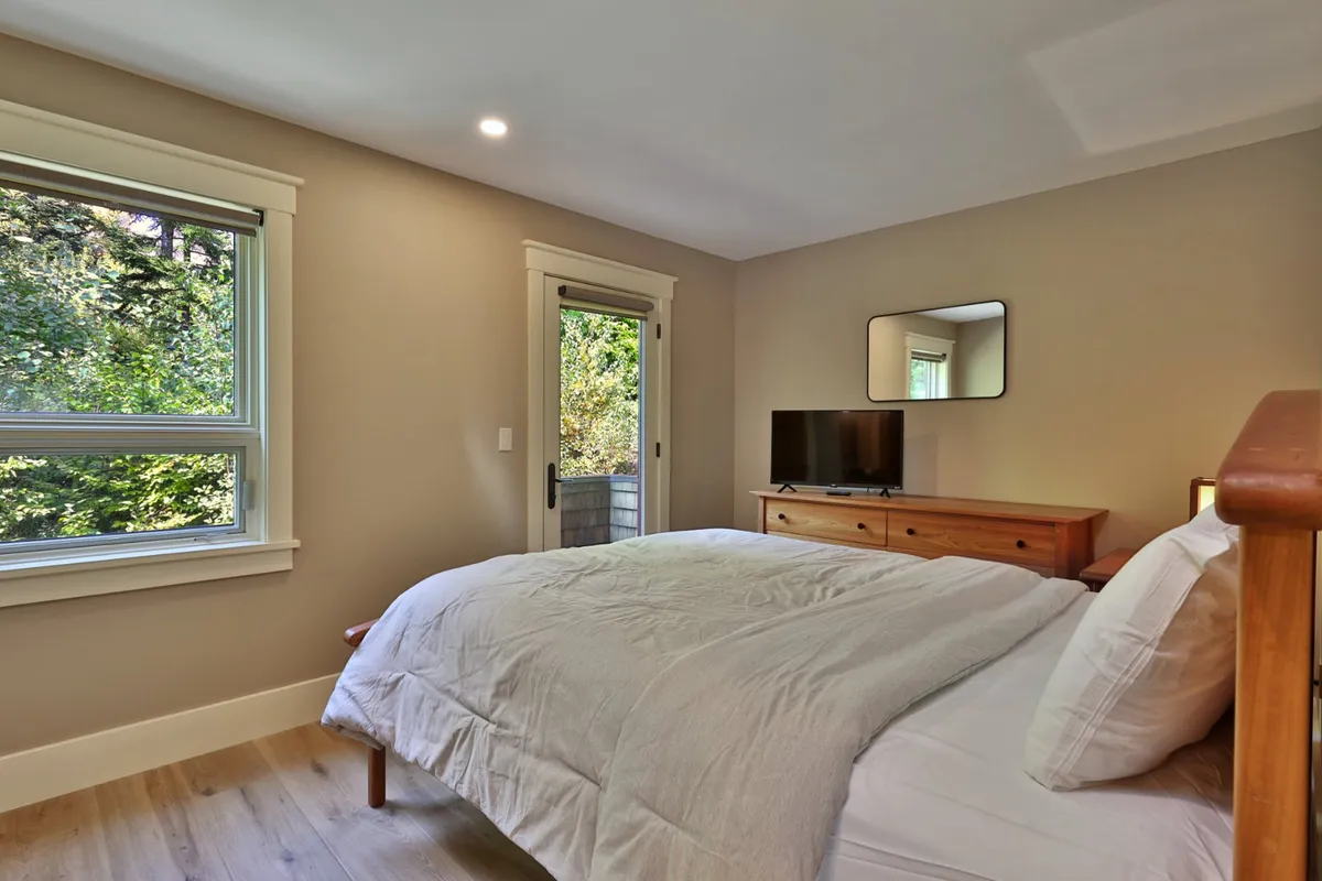 A tranquil bedroom featuring a cozy bed, soft linens, a window with serene views of nature, a dresser with a TV, and a mirror. The room is bathed in natural light, emphasizing restful comfort and a peaceful atmosphere.