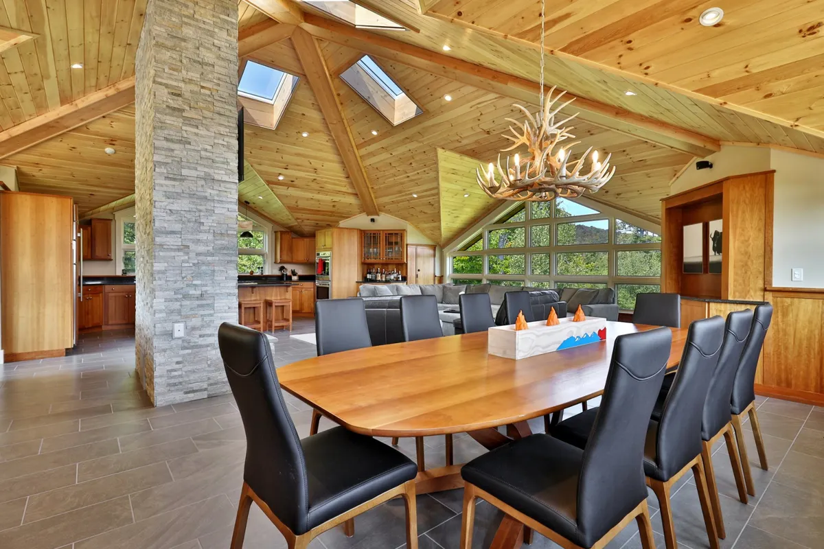 The photo shows a large dining table set beneath an antler chandelier, with a stone pillar and skylights providing a cozy yet communal atmosphere. This setting is perfect for group dinners or celebrations after a day on the slopes.