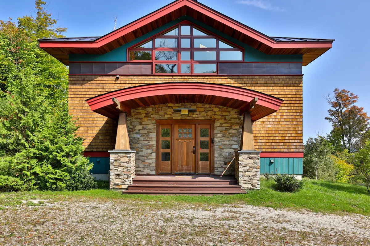 This exterior image features a welcoming wooden front door flanked by stone pillars and a red roof, with large windows inviting views both in and out. The setting includes a porch area ideal for gathering and open space for entertaining guests or enjoying the surrounding nature.