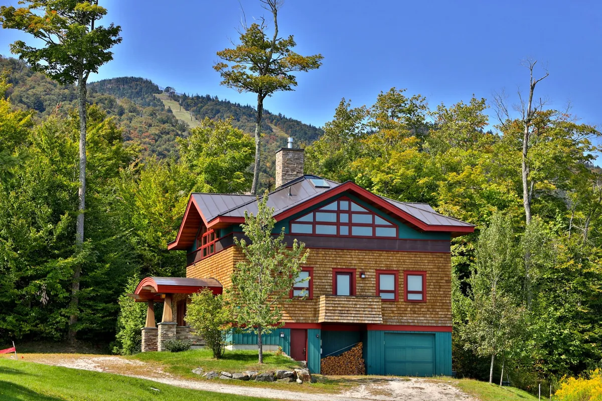 This exterior image features the house nestled among trees with a striking mountain backdrop, conveying immediate proximity to the slopes and the natural beauty of Killington and Bear Mountain. The porch and outdoor spaces are visible, reinforcing the idea of direct outdoor adventure access.