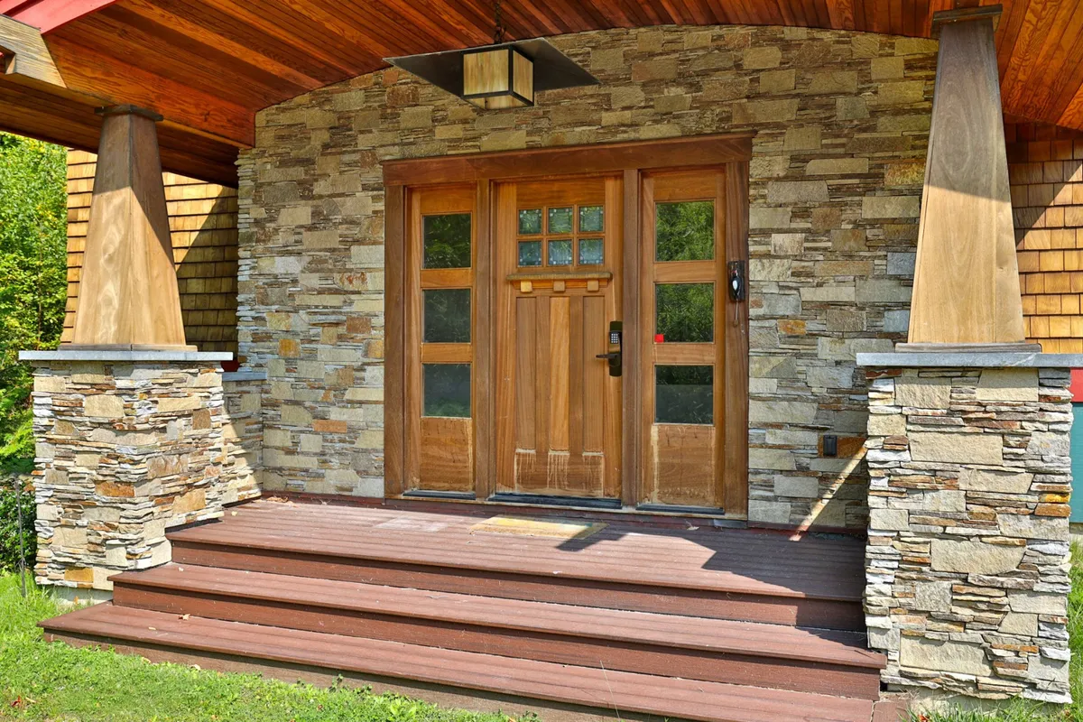 A welcoming exterior entrance with wooden double doors, sturdy stone columns, and an overhanging roof sets a warm first impression. The space suggests family members or guests arriving home or gathering at the threshold.