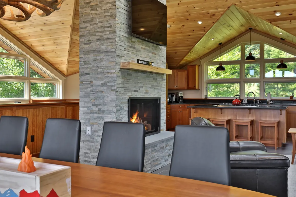 This picture showcases an open-concept living and dining area, with a stone fireplace as a central feature, large windows offering scenic views, a dining table ready for gatherings, and a visible kitchen area in the background. Warm wood tones and elegant finishes set the mood for family feasts and social events.